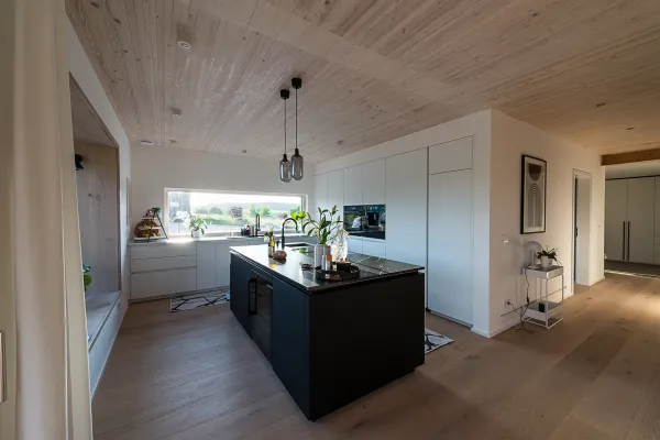 Moderne weiße Küche mit schwarzem Küchenblock in der Mitte. Oben zu sehen die Holzdecke.